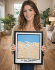 Happy woman displaying framed San Juan Puerto Rico 3D wooden city map, precision laser-cut street design with natural wood layers and blue sea detail, handmade wall decor in cozy living room.