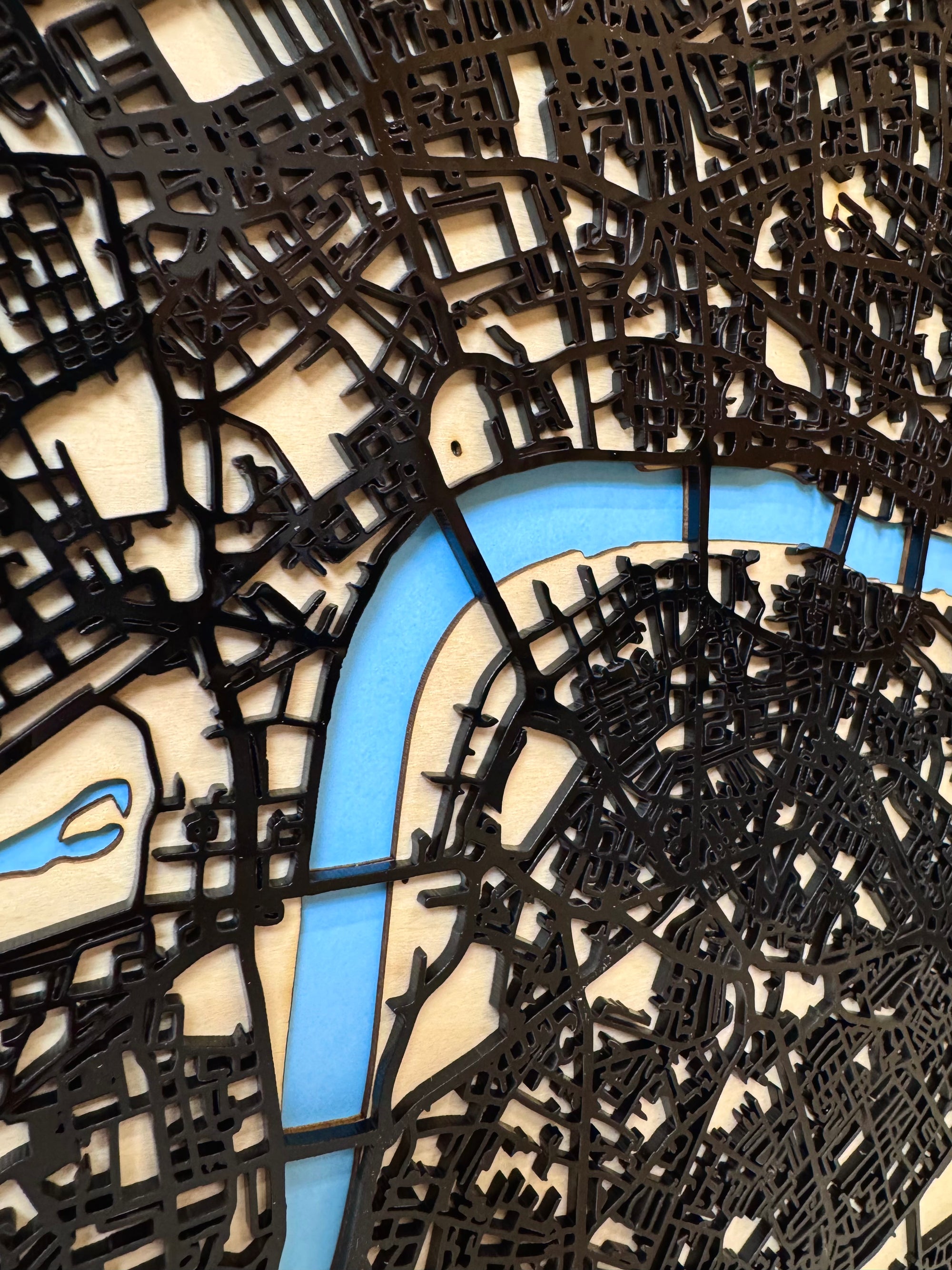 Close-up London 3D street map wall art in natural wood frame – precision laser-cut black layered road network mounted over light wood backing with blue acrylic River Thames detail, handcrafted modern home decor made in France.