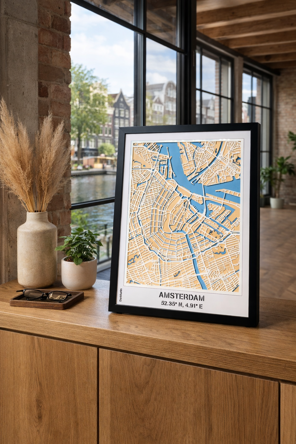 Amsterdam city map wall art frame displayed on a console table in a modern loft living room with a view over Amsterdam canals – handcrafted laser-cut wooden city map with blue waterways and detailed streets for elegant home decor