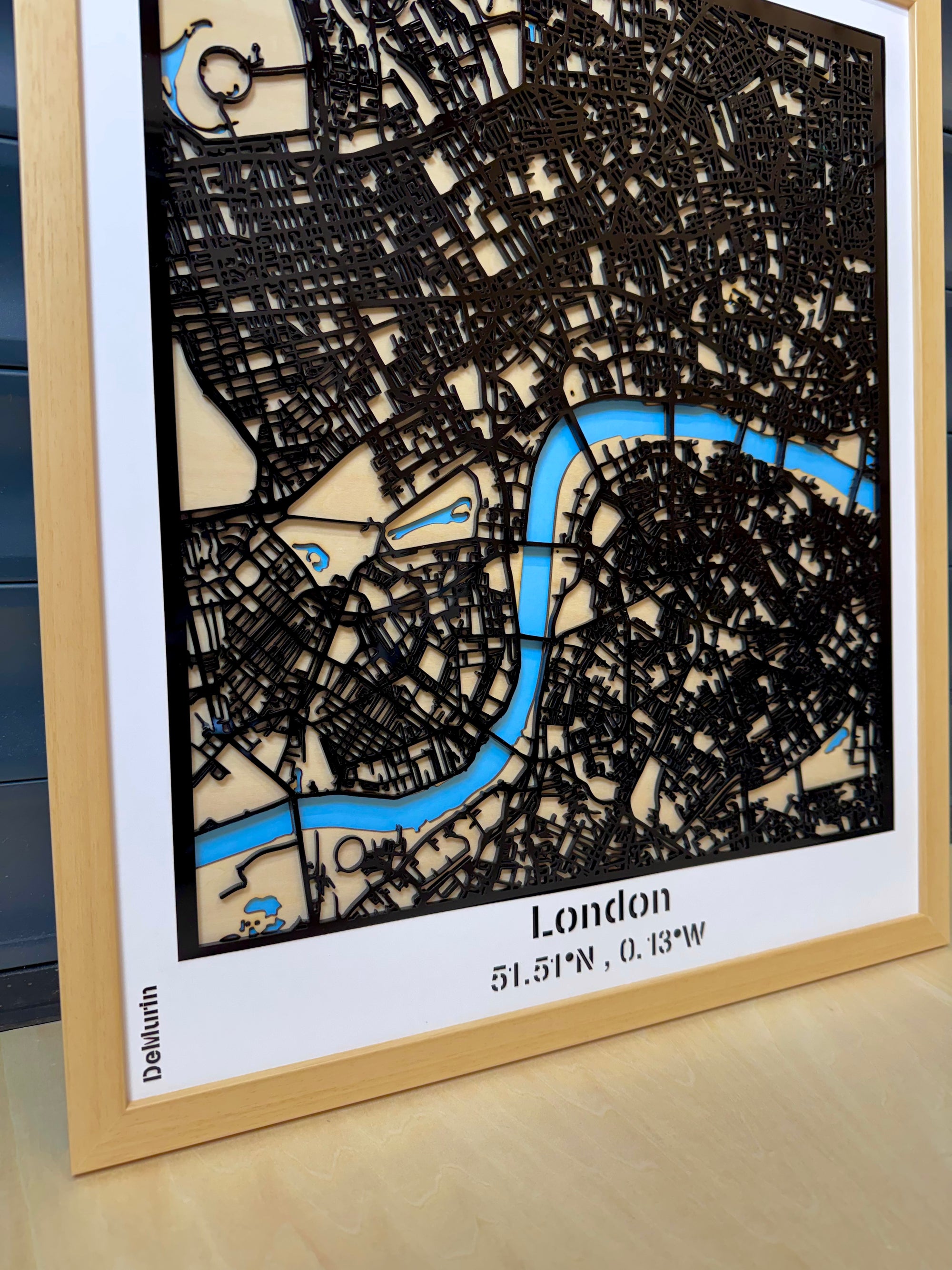 London city map wall decor in natural oak wood frame displayed in a contemporary living room – multi-layer 3D laser-cut street map with intricate road details, River Thames in blue acrylic, handcrafted modern wall art made in France.
