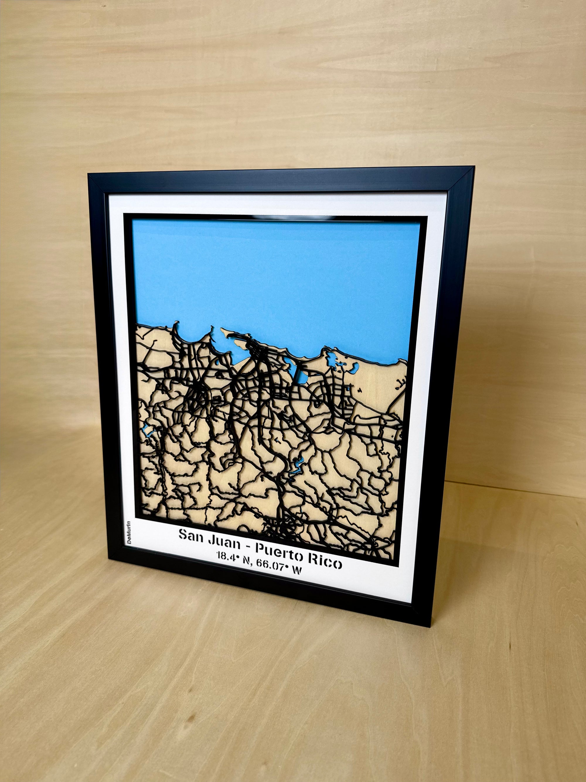 Angled three-quarter view of San Juan Puerto Rico framed 3D city map featuring white precision laser-cut street network, natural wood layers, blue sea detail and engraved coordinates, handmade wall decor.