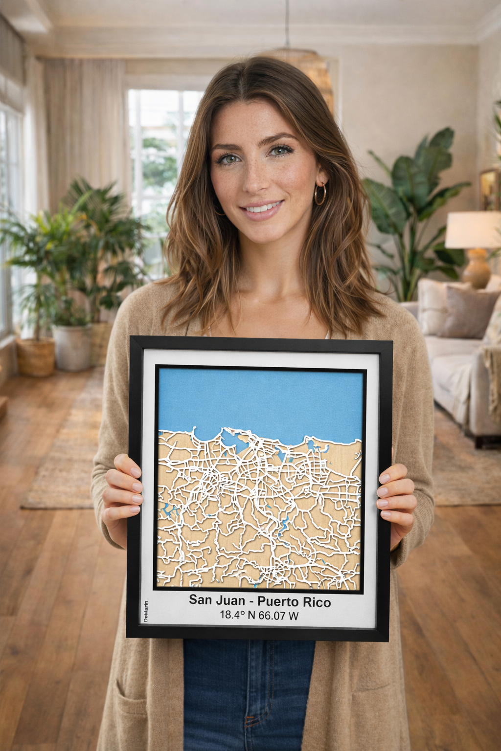 Happy woman displaying framed San Juan Puerto Rico 3D wooden city map, precision laser-cut street design with natural wood layers and blue sea detail, handmade wall decor in cozy living room.